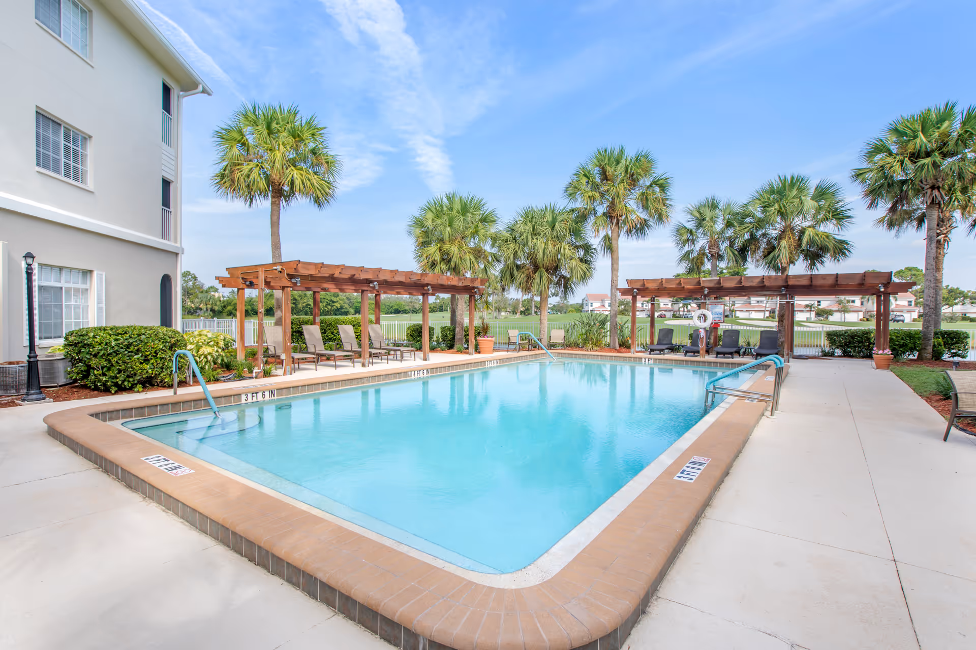 Outdoor swimming pool area at Brookdale Fort Myers Cypress Lake with clear blue water, surrounded by a concrete deck. There are wooden pergolas with lounge chairs underneath, palm trees around the pool, and a building visible on the left side. The sky is clear with some clouds.