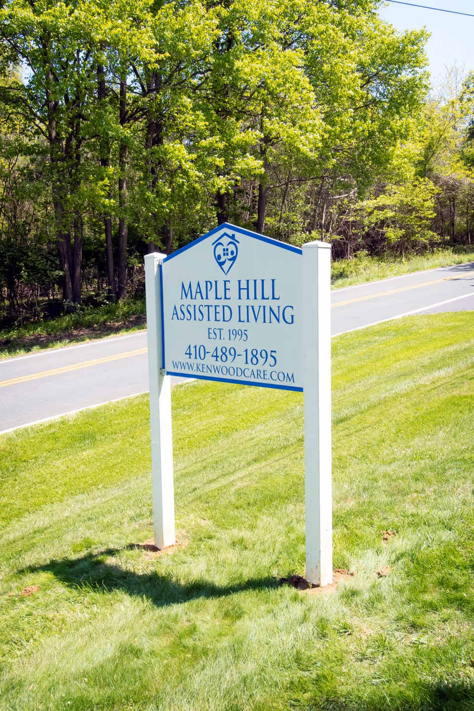 A white and blue sign for Maple Hill Assisted Living standing on a grassy area next to a road, with green trees in the background. The sign includes the establishment year 1995, a phone number, and a website URL.