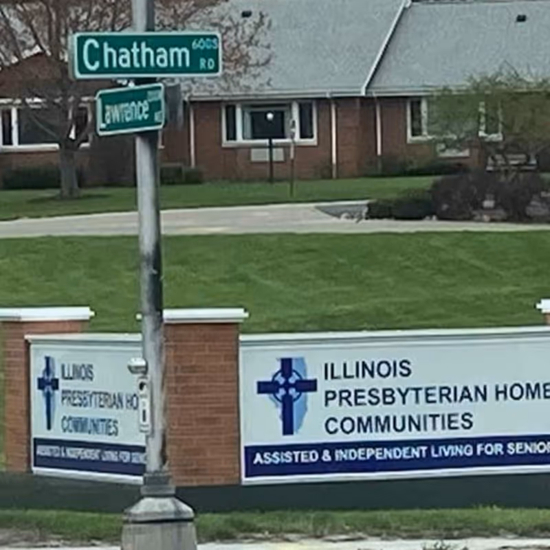 Outdoor view of a street corner with a street sign reading 'Chatham 6605 Rd' and 'Entrance'. Below the street signs, there is a large sign for Illinois Presbyterian Home Communities indicating assisted and independent living for seniors. The sign features a blue cross logo and is set against a background of grass and a brick building.