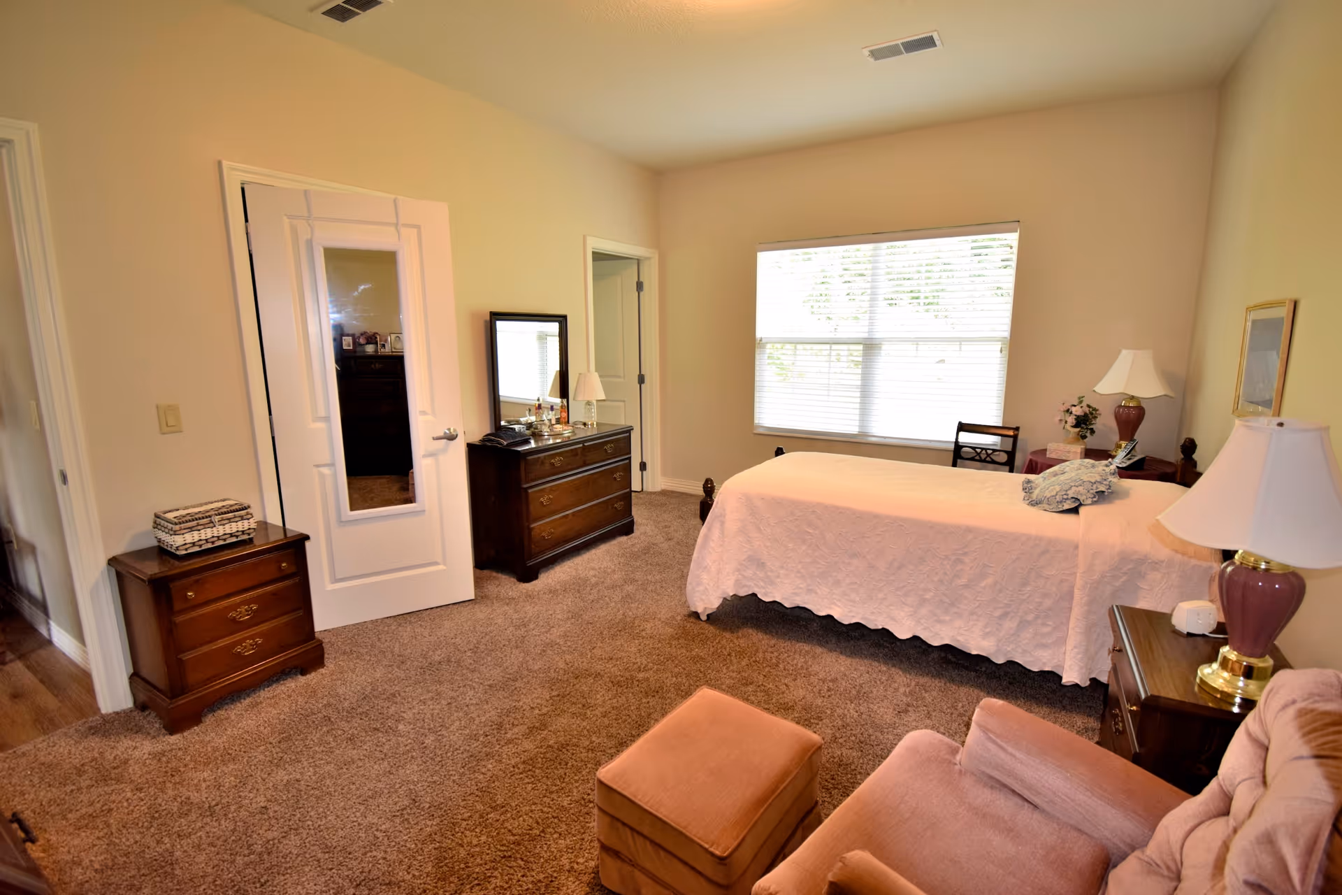 A cozy bedroom with beige walls and carpeted floor featuring a single bed with a white quilt, a wooden dresser with a mirror, a nightstand with a lamp, a cushioned armchair with an ottoman, and a window with blinds letting in natural light.