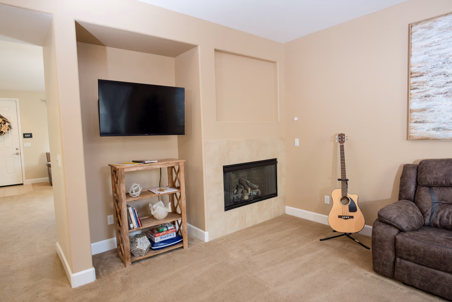 A cozy living room area with beige walls and carpet. A flat-screen TV is mounted on the wall above a wooden shelf holding books, decorative items, and board games. There is a tiled fireplace next to the shelf. A guitar on a stand is placed near a dark brown recliner sofa. A large abstract painting hangs on the wall above the sofa. In the background, a hallway leads to a door with an exit sign.