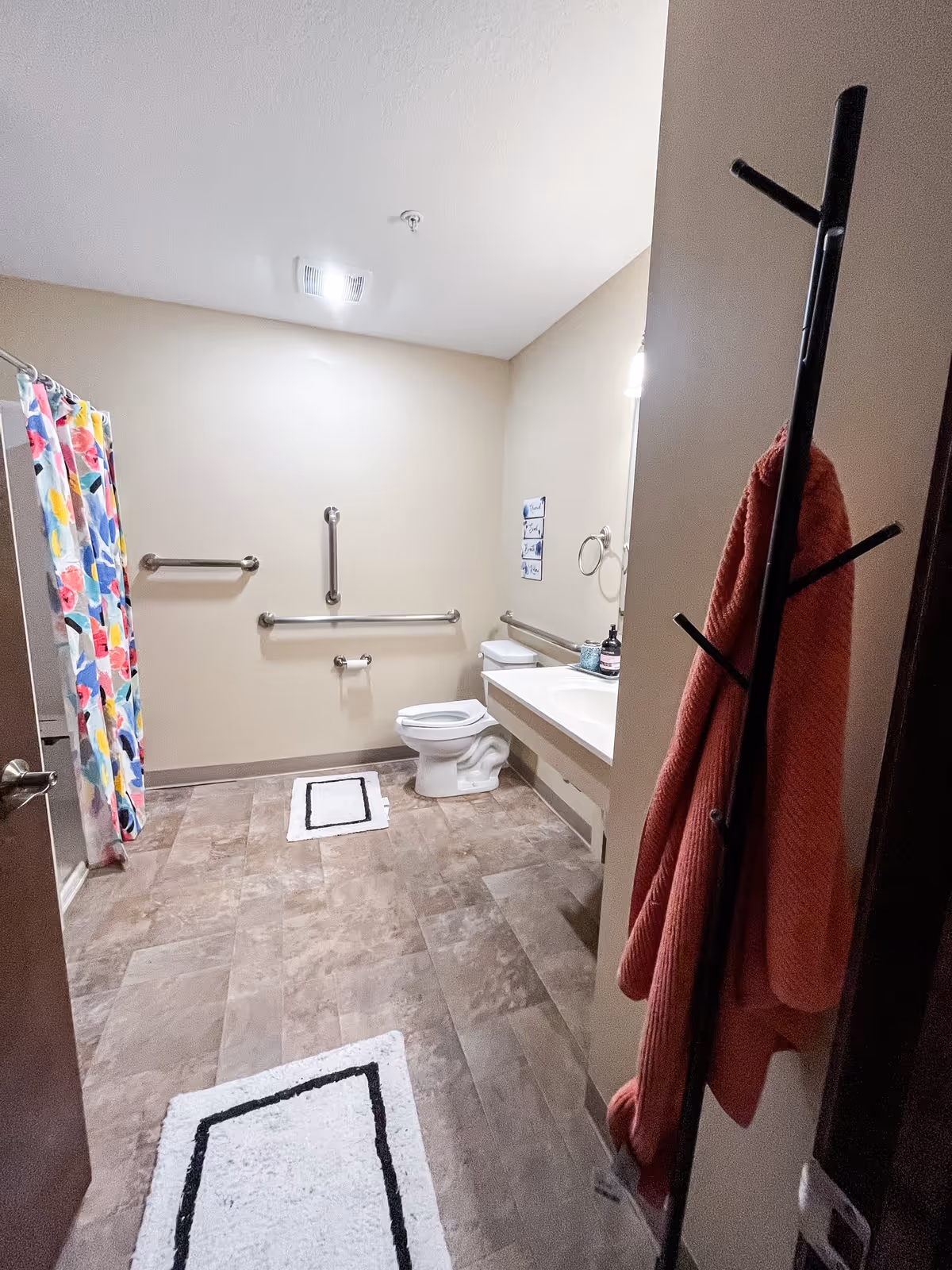A spacious bathroom with beige walls and tiled floor. There is a toilet with grab bars on the wall behind and beside it, a sink with a countertop and a mirror above it, and a colorful floral shower curtain on the left side. Two white bath mats with black borders are on the floor. A red towel hangs on a black coat rack near the entrance.