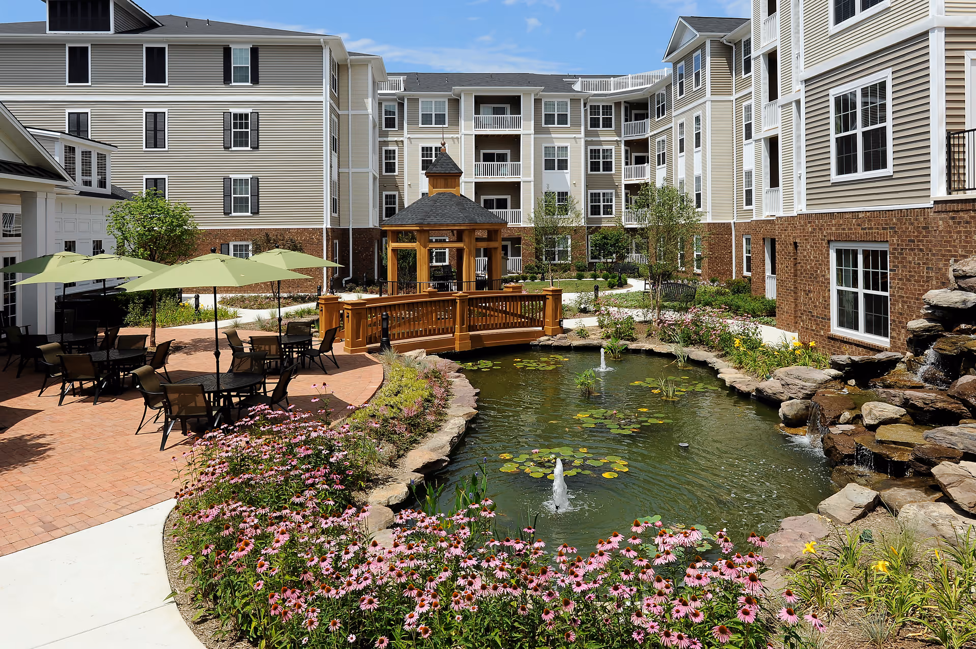 Outdoor courtyard of Regency Crest senior living facility featuring a pond with water fountains and lily pads, surrounded by flowering plants and rocks. There is a wooden gazebo and a small wooden bridge over the pond. Patio area with tables, chairs, and green umbrellas is visible on the left side. The multi-story building with beige siding and brick accents surrounds the courtyard under a blue sky with some clouds.