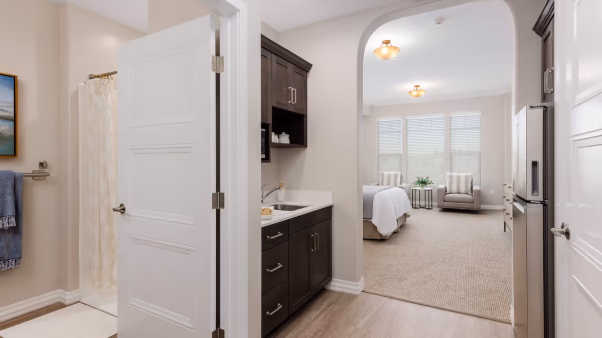 Interior view of a senior studio showing a kitchenette and open bathroom on the left with a bed and seating area visible beyond an arched doorway.