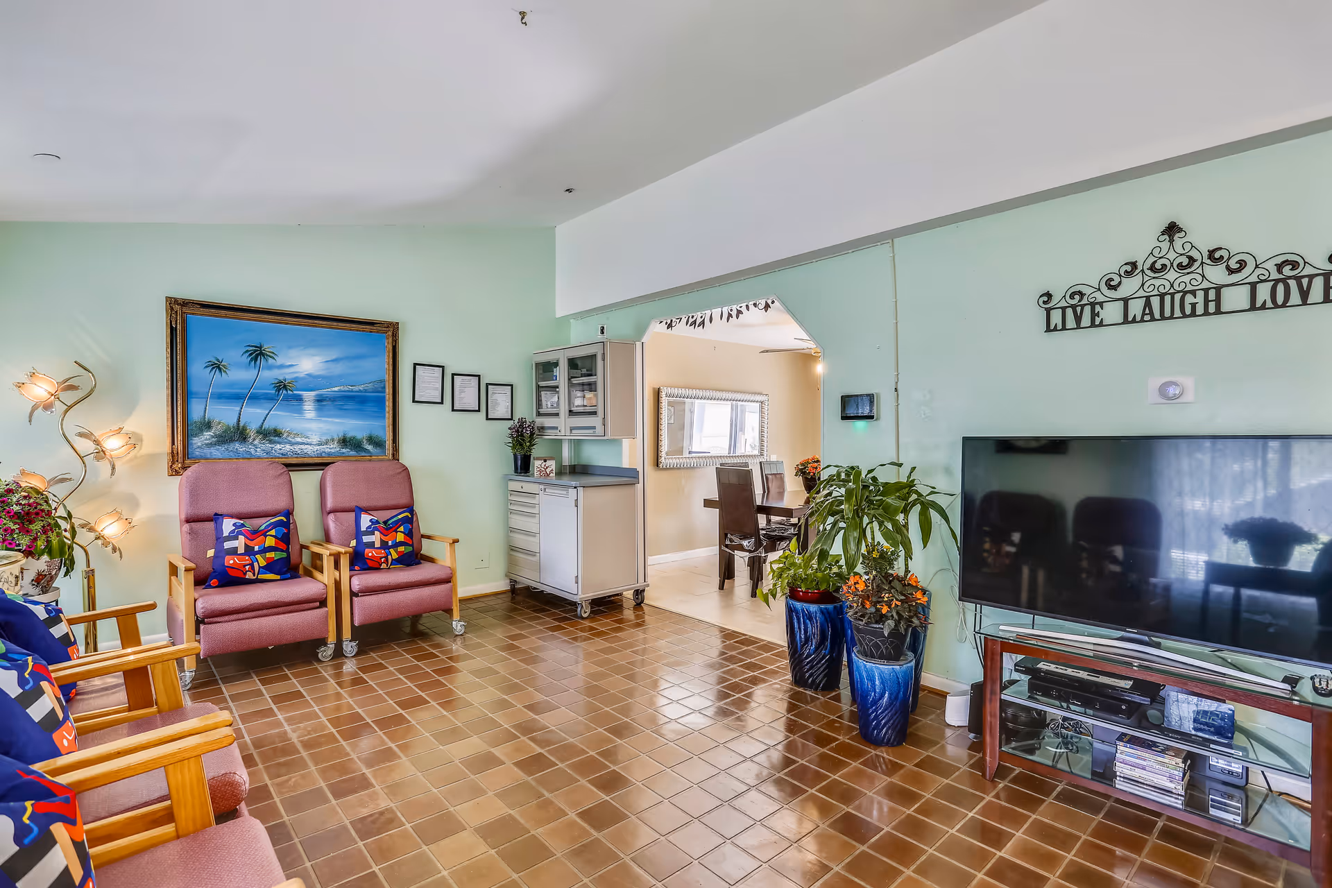 A living room area with light green walls and brown tiled floor. There are four pink cushioned chairs with colorful pillows arranged along the left side. A floor lamp with multiple lights shaped like flowers stands next to the chairs. A large painting of a beach scene with palm trees hangs on the wall. On the right side, there is a flat-screen TV on a glass stand with various electronic devices and DVDs below. Two large blue plant pots with green and orange plants are placed near the TV. Above the TV, a decorative wall sign reads 'LIVE LAUGH LOVE'. An open doorway leads to a dining area with a table and chairs, and a large mirror on the wall.