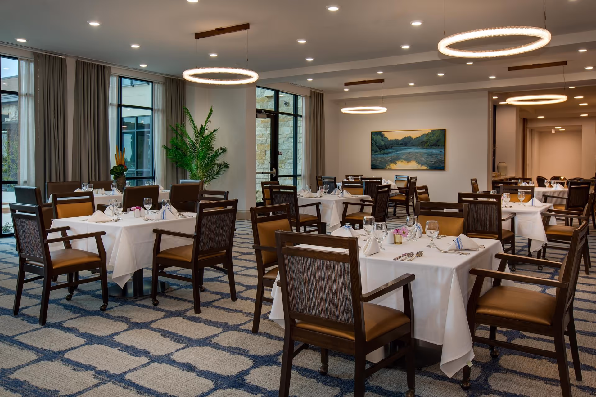 Spacious dining room with multiple tables set with white tablecloths, chairs, and circular pendant lights.