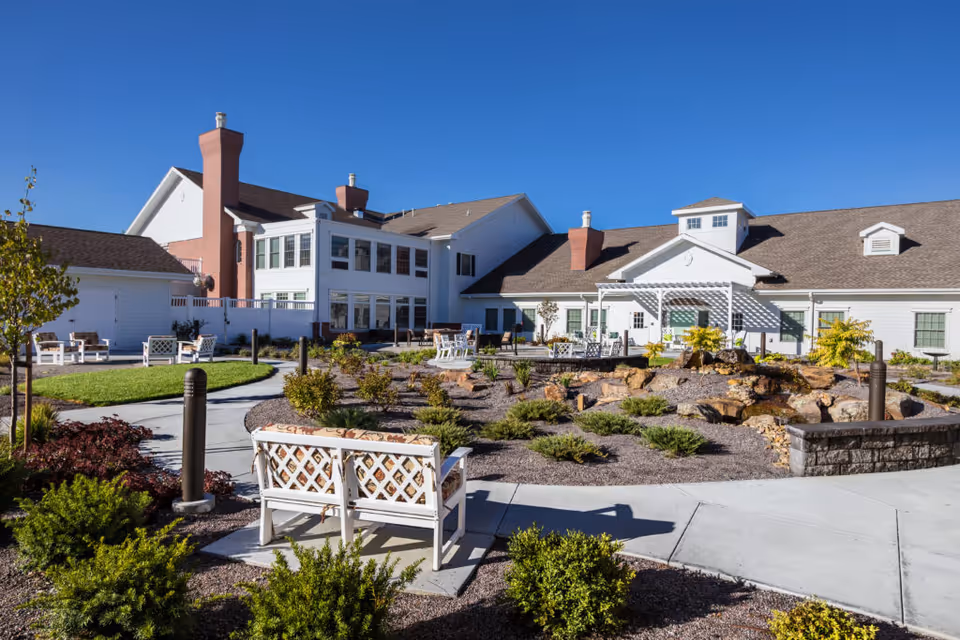 Outdoor patio area of CountryHouse Residence featuring white benches with cushions, landscaped garden beds with shrubs and rocks, paved walkways, and a large white building with multiple chimneys and windows under a clear blue sky.