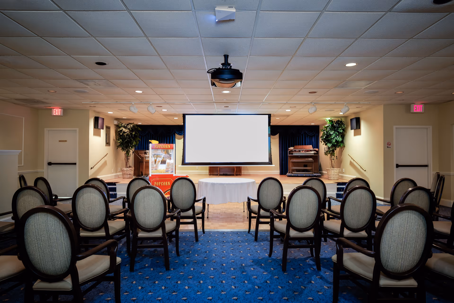 A small auditorium or meeting room with rows of upholstered chairs facing a stage. On the stage, there is a white projection screen, a popcorn machine, a round table covered with a white tablecloth, and two potted plants on either side. The room has a blue carpet with a patterned design and a drop ceiling with recessed lighting.