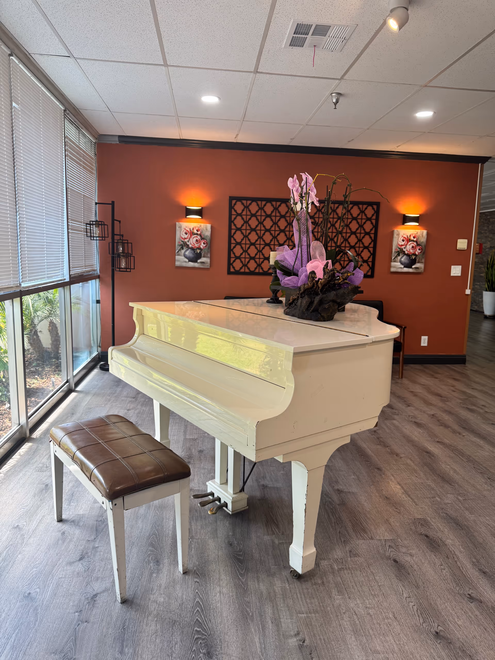 White grand piano with a bench and floral arrangement in a bright lounge area with large windows and a red accent wall.