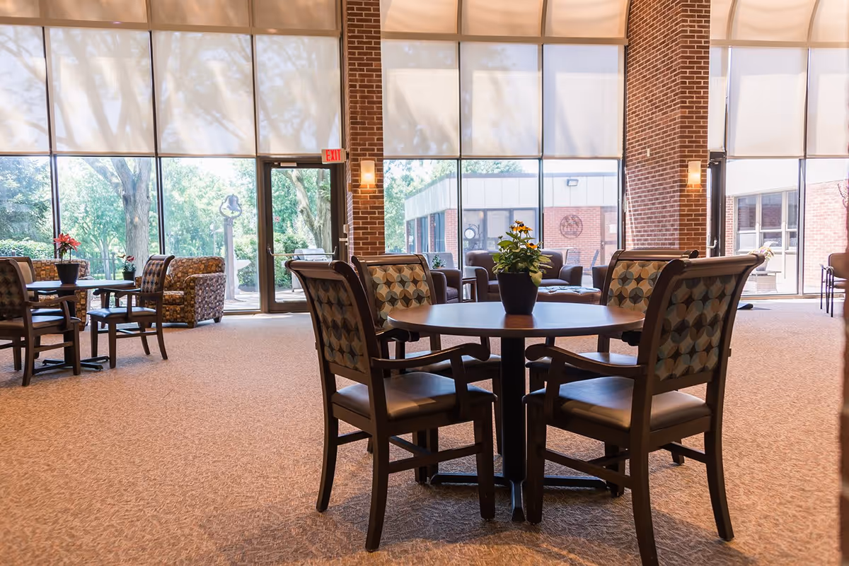 A bright and spacious common area in a senior living facility with large floor-to-ceiling windows letting in natural light. The room features round wooden tables with patterned cushioned chairs and small potted plants on the tables. There are additional seating areas with armchairs near the windows, and the walls are made of exposed brick.