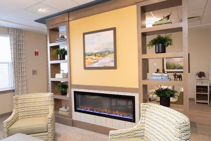 A cozy living room area in a senior living facility featuring a modern electric fireplace set in a yellow accent wall with built-in wooden shelves on either side. The shelves hold decorative items including potted plants, books, and small sculptures. Two patterned armchairs are positioned facing the fireplace. A window with curtains is visible on the left side, and a framed landscape painting hangs above the fireplace.