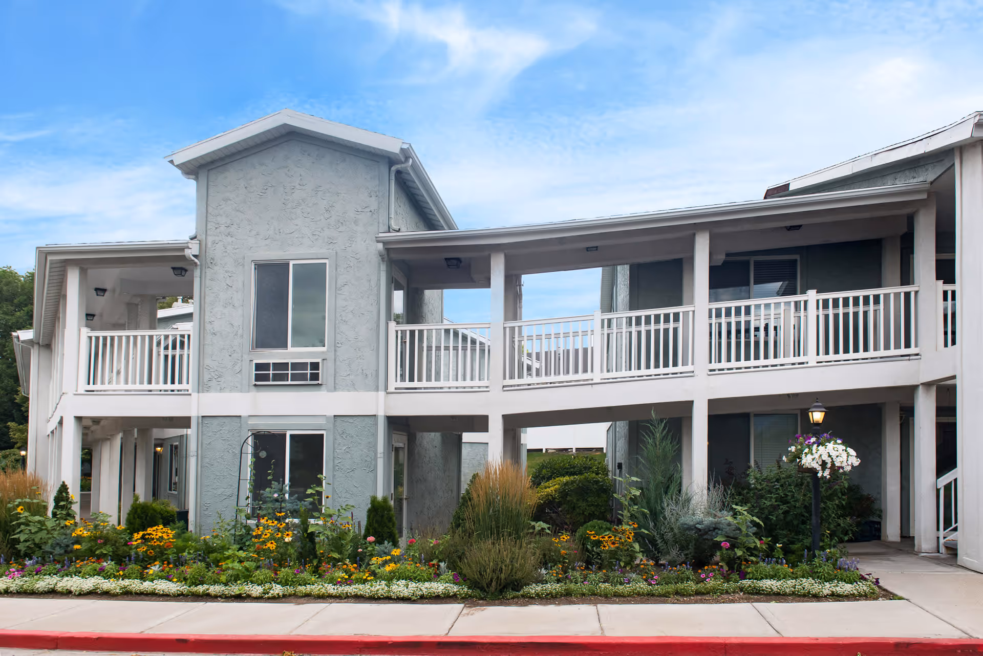 Exterior view of a two-story building with a light gray stucco finish and white railings. The building has a covered walkway on the second floor and a landscaped garden with colorful flowers and greenery in front. The sky is partly cloudy with blue tones.