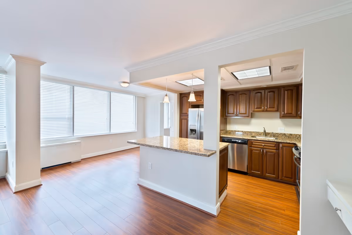 Bright and spacious interior view of an apartment with large windows covered by blinds, wooden flooring, and an open kitchen featuring dark wooden cabinets, granite countertops, stainless steel appliances, and pendant lighting.