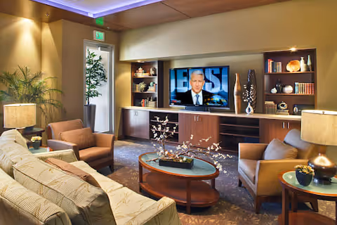 A cozy living room with a beige patterned sofa, two brown armchairs, a round glass coffee table with a decorative centerpiece, and a wooden entertainment center with a flat-screen TV displaying a man speaking. The room is warmly lit with table lamps and recessed ceiling lights, and there are shelves with books and decorative items. A potted plant is near a door with an exit sign above it.