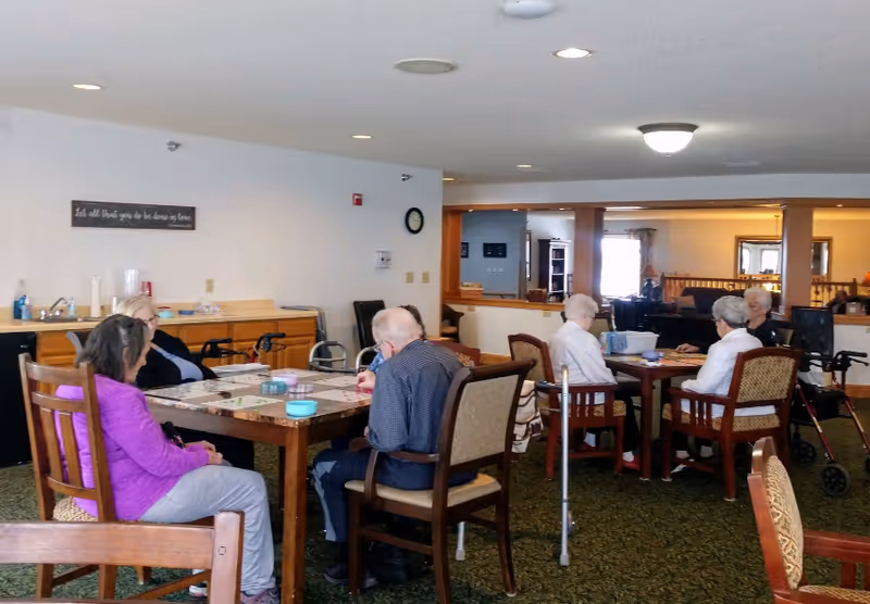Several elderly residents seated around tables in a communal dining/activity room.