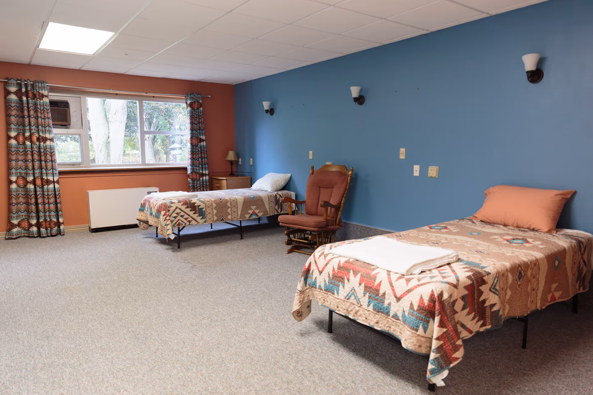 A spacious senior bedroom with two twin beds covered in patterned quilts, a rocking chair, nightstand and a window against blue and terracotta-colored walls.