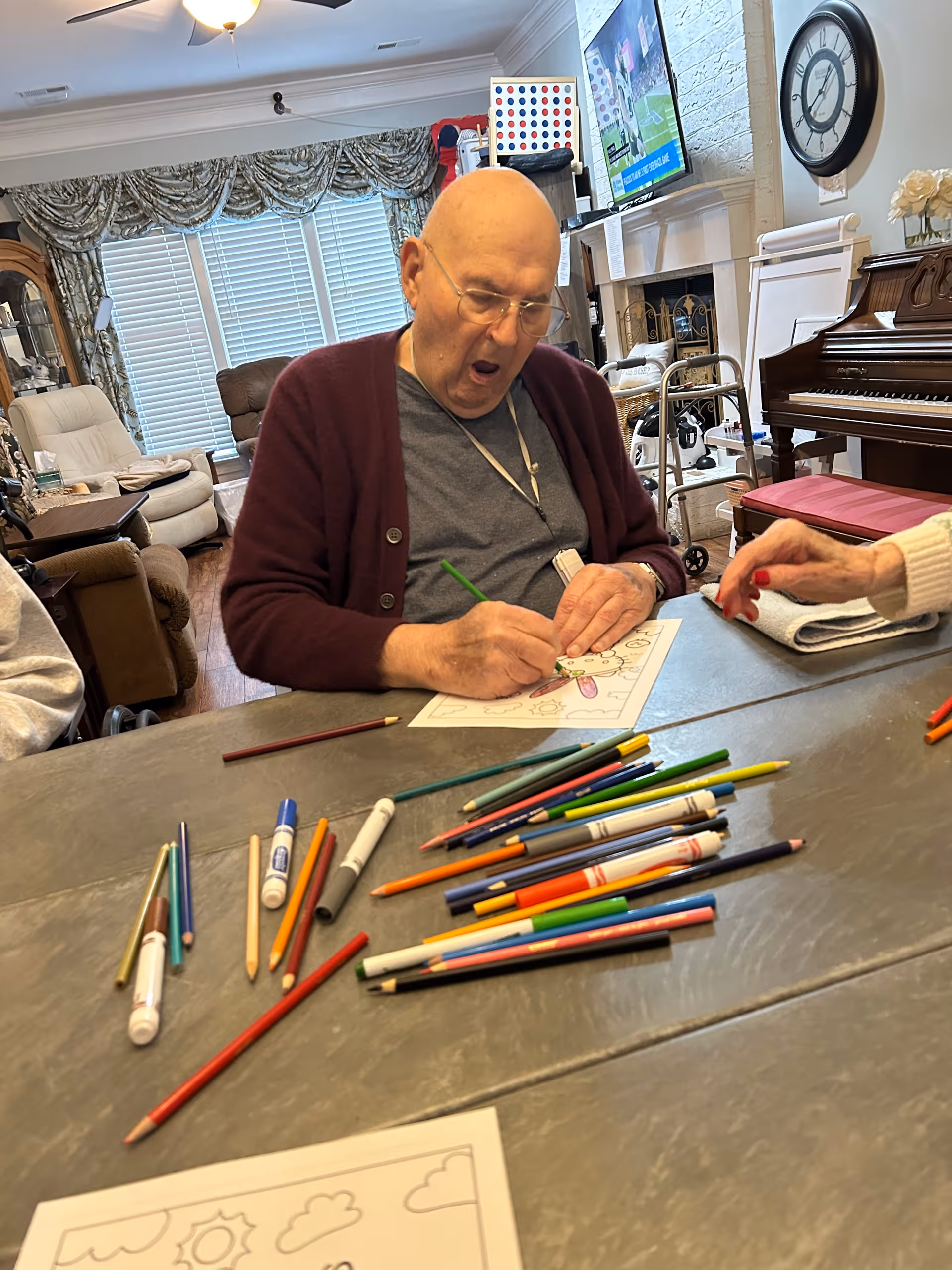 An elderly man wearing glasses and a maroon cardigan is sitting at a table in a living room, coloring a picture with colored pencils and markers scattered on the table. Another person's hand is visible on the right side of the table. The room has a piano, a clock on the wall, a fireplace, and large windows with curtains.