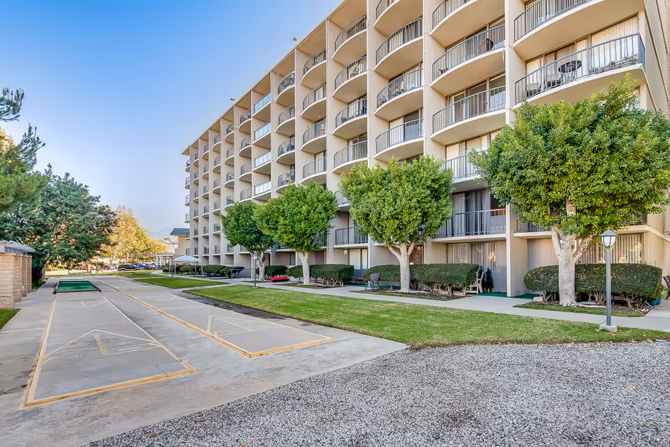 Exterior view of a multi-story senior living building with balconies, landscaped grounds and outdoor shuffleboard courts.
