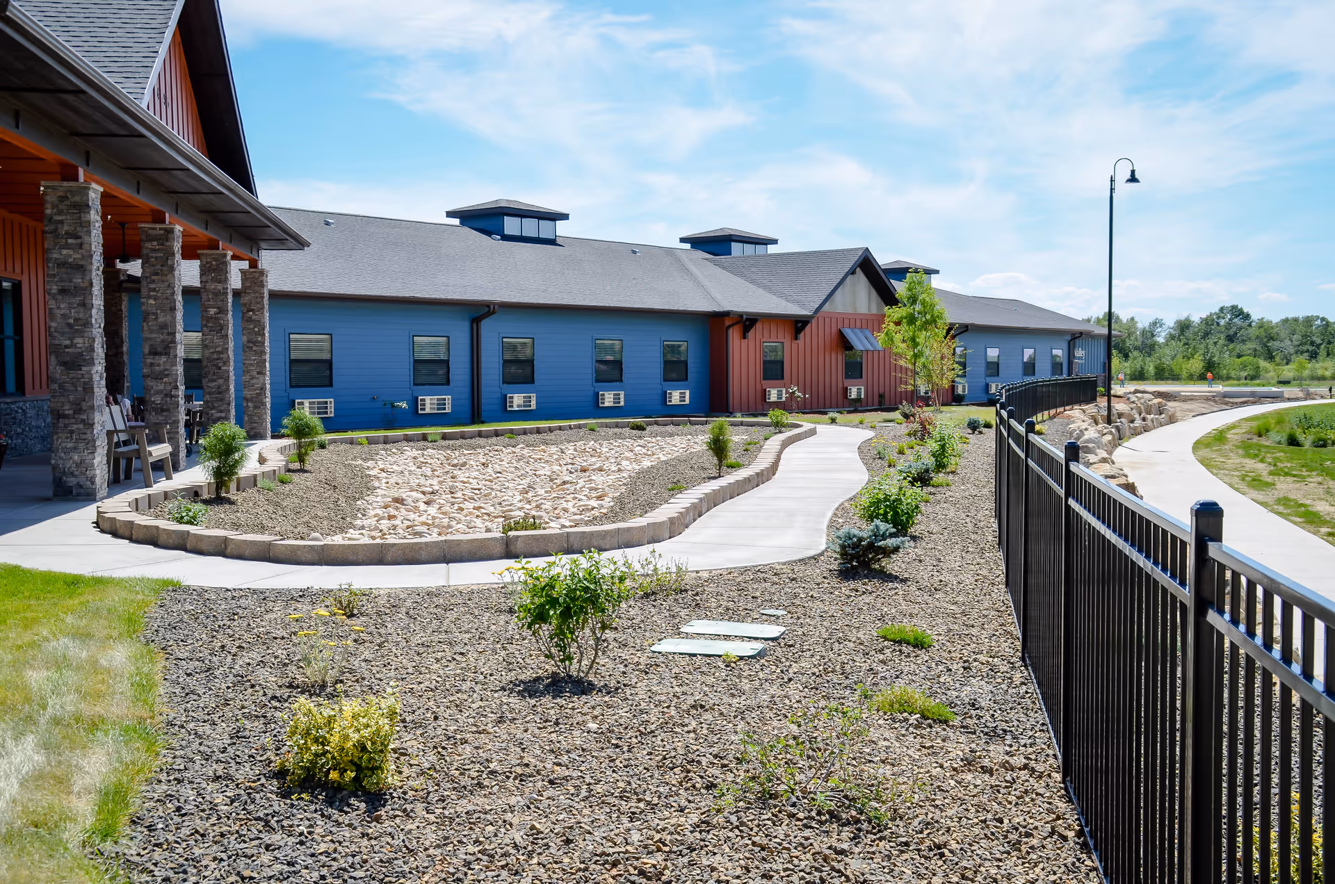 Exterior view of a senior living facility with a blue and red building, a paved walkway, landscaped garden beds with small plants and shrubs, a black metal fence, and a clear blue sky.
