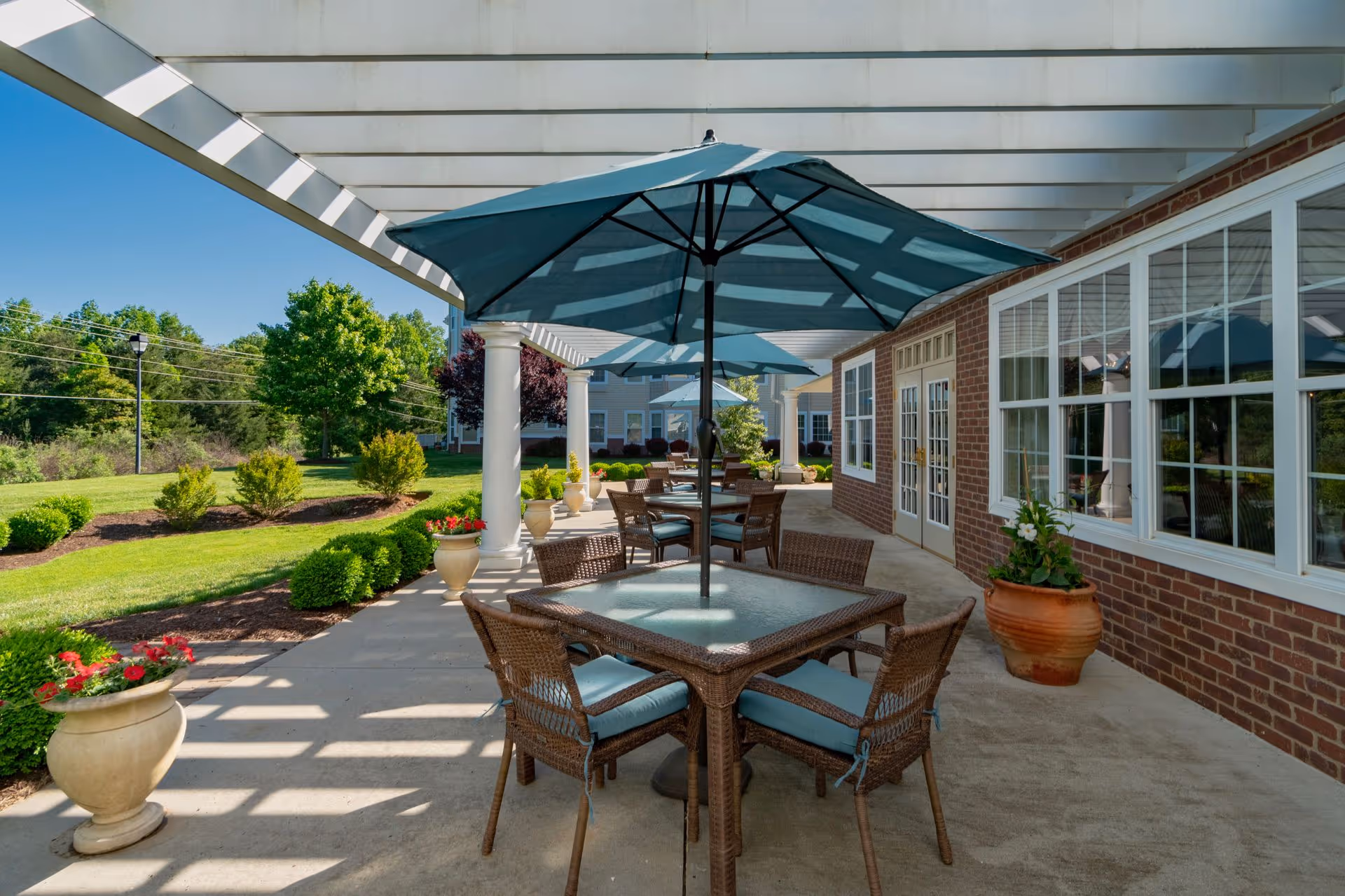Outdoor covered patio with wicker tables, blue umbrellas, and potted plants beside a brick building and landscaped lawn.