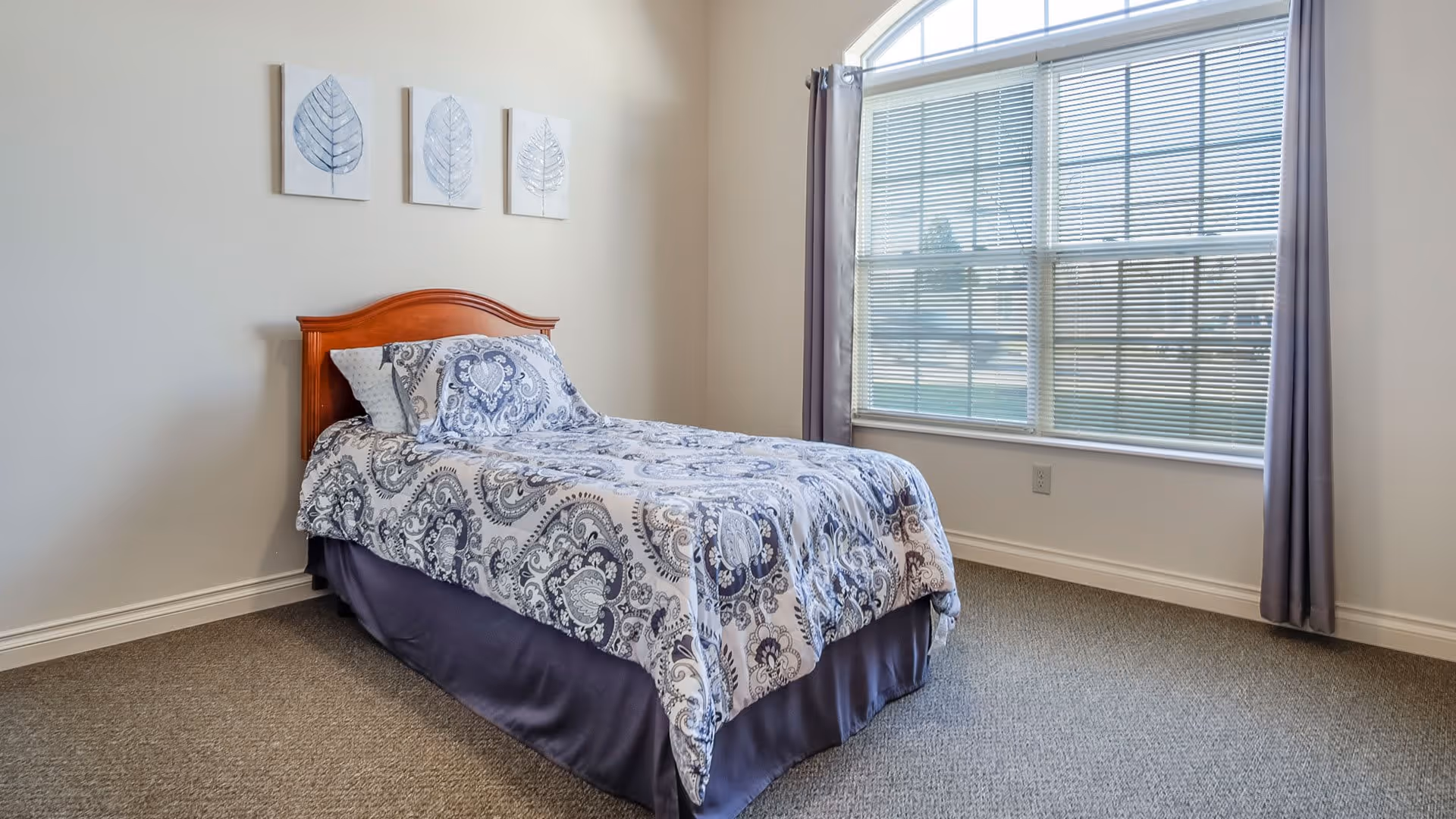A simple bedroom with a single bed featuring a wooden headboard and blue patterned bedding. The room has beige walls, a large window with blinds and gray curtains, and three framed leaf artworks hanging above the bed.