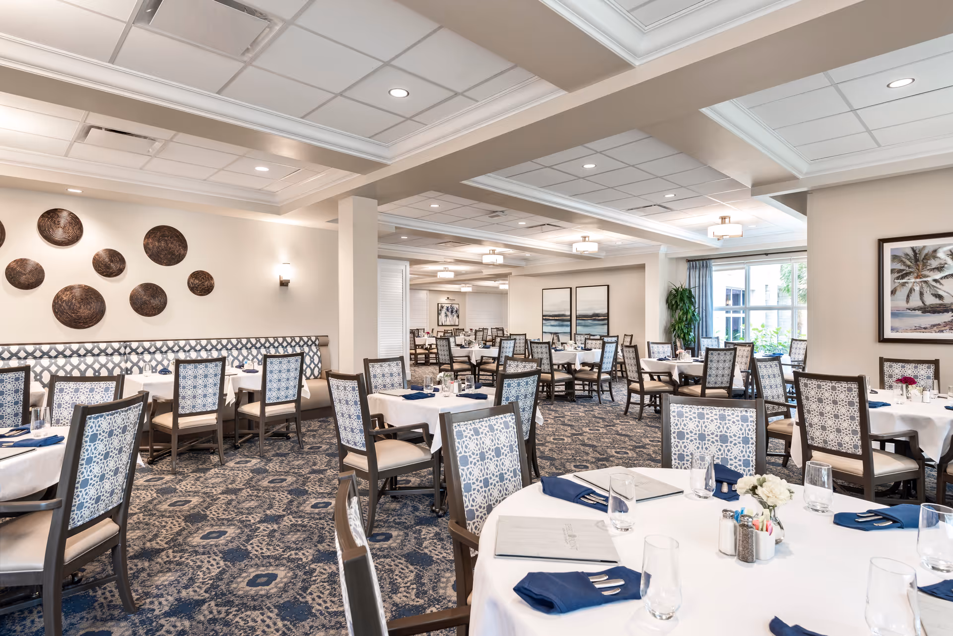 A spacious and well-lit dining room with multiple round and rectangular tables covered with white tablecloths. Each table is set with glassware, silverware wrapped in navy blue napkins, and small flower arrangements. The chairs have patterned upholstery, and the room features decorative wall art, a patterned carpet, and large windows allowing natural light to enter.