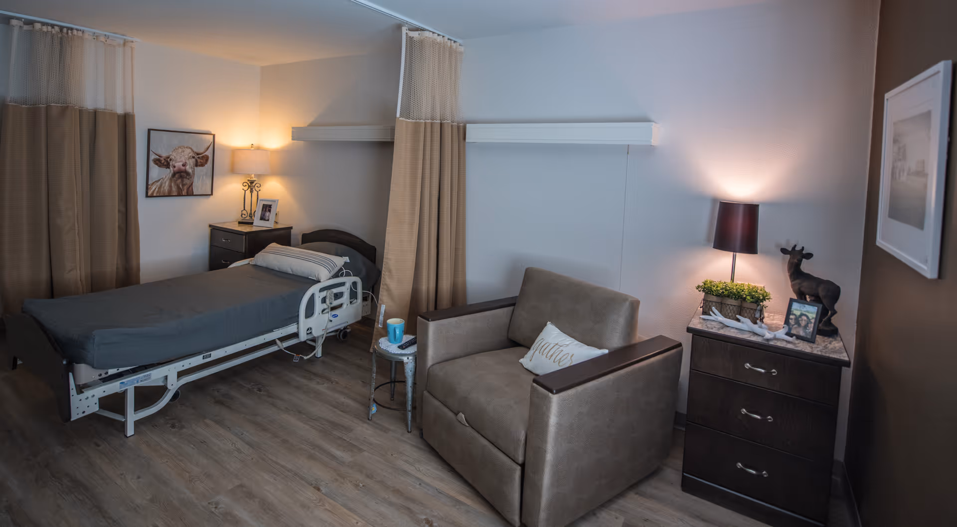 A cozy room in a healthcare facility featuring a single hospital bed with gray bedding, a small bedside table with a lamp and framed photo, a comfortable armchair with a pillow, and a dark wooden dresser with a lamp, decorative plants, and a small animal figurine. The room has wood flooring, beige curtains, and framed artwork on the walls.