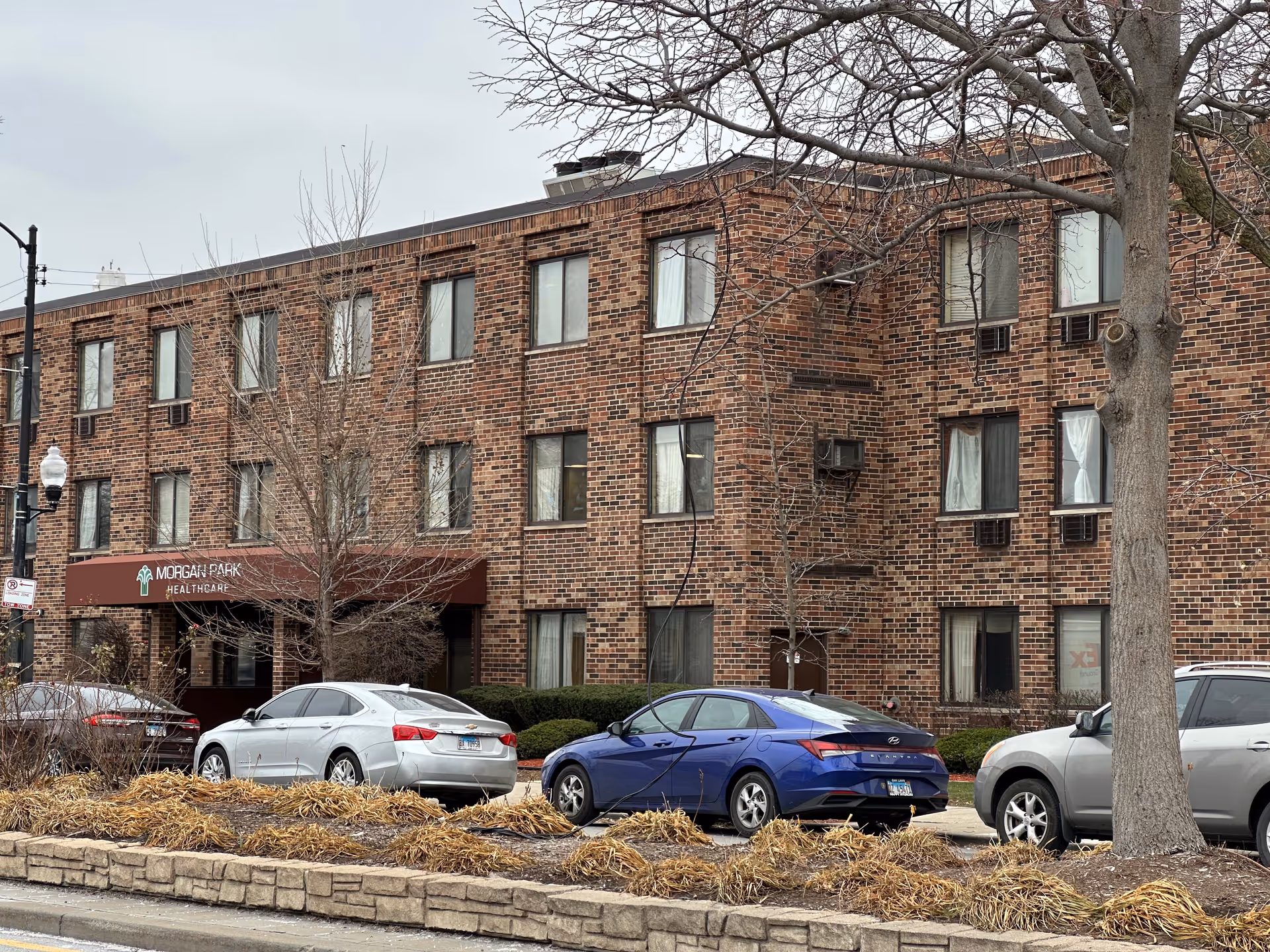 Exterior view of a three-story brick building with multiple windows and a maroon awning that reads 'Morgan Park Healthcare'. Several cars are parked in front of the building, and leafless trees and dry landscaping are visible along the sidewalk.