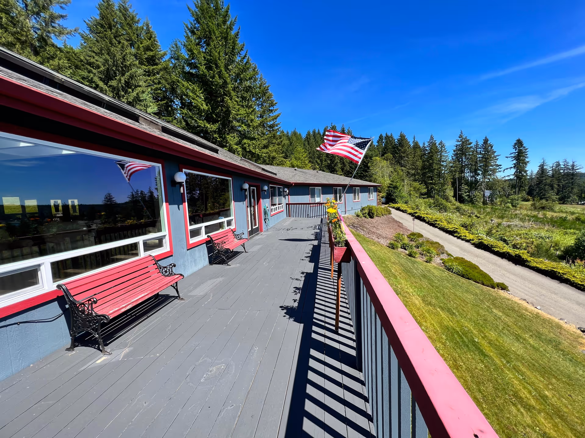 Long outdoor deck of an assisted living building with benches, American flags, railing and landscaped grounds under a clear blue sky.
