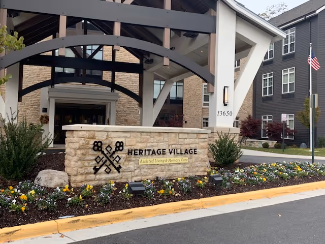 Stone sign reading 'Heritage Village Assisted Living & Memory Care' sits amid landscaping in front of the facility's covered entrance and building.