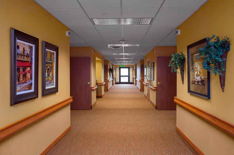A long, well-lit hallway in a senior living facility with beige walls and carpeted floor. The walls are decorated with framed paintings and wall-mounted plants. Several closed wooden doors line both sides of the hallway, and a glass exit door is visible at the far end.
