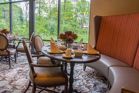 Sunlit dining area with a set round table, chairs and a curved upholstered banquette beside large windows overlooking greenery.