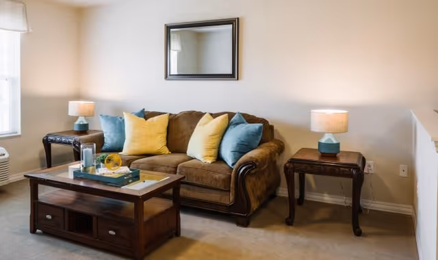 A cozy living room with a brown sofa adorned with yellow and blue throw pillows, a wooden coffee table with decorative items, two side tables each with a lamp, a window with white curtains, and a rectangular mirror hanging on the wall above the sofa.