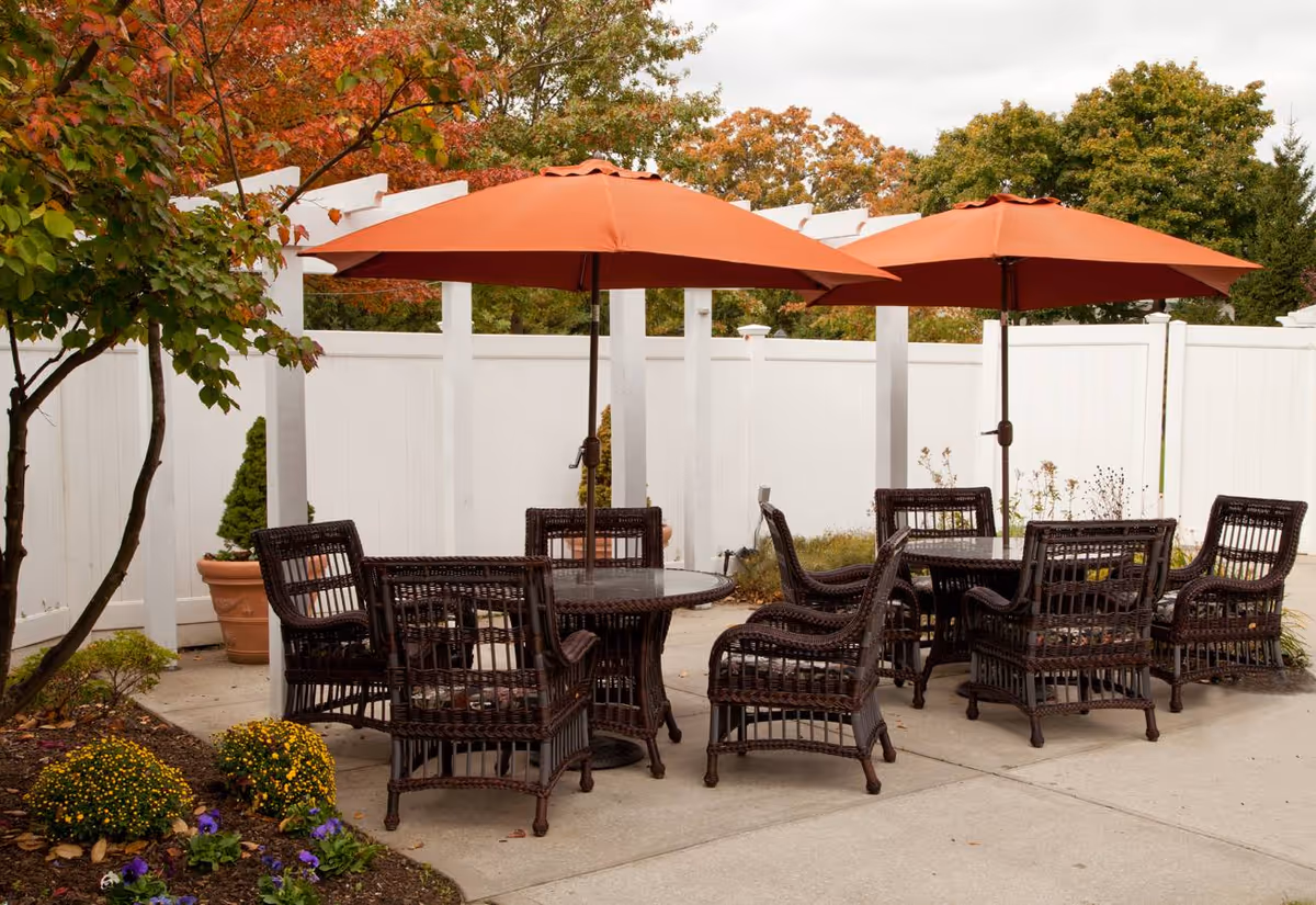 Outdoor patio area with two round tables surrounded by wicker chairs and shaded by large orange umbrellas. The space is enclosed by a white fence and decorated with potted plants and autumn foliage.