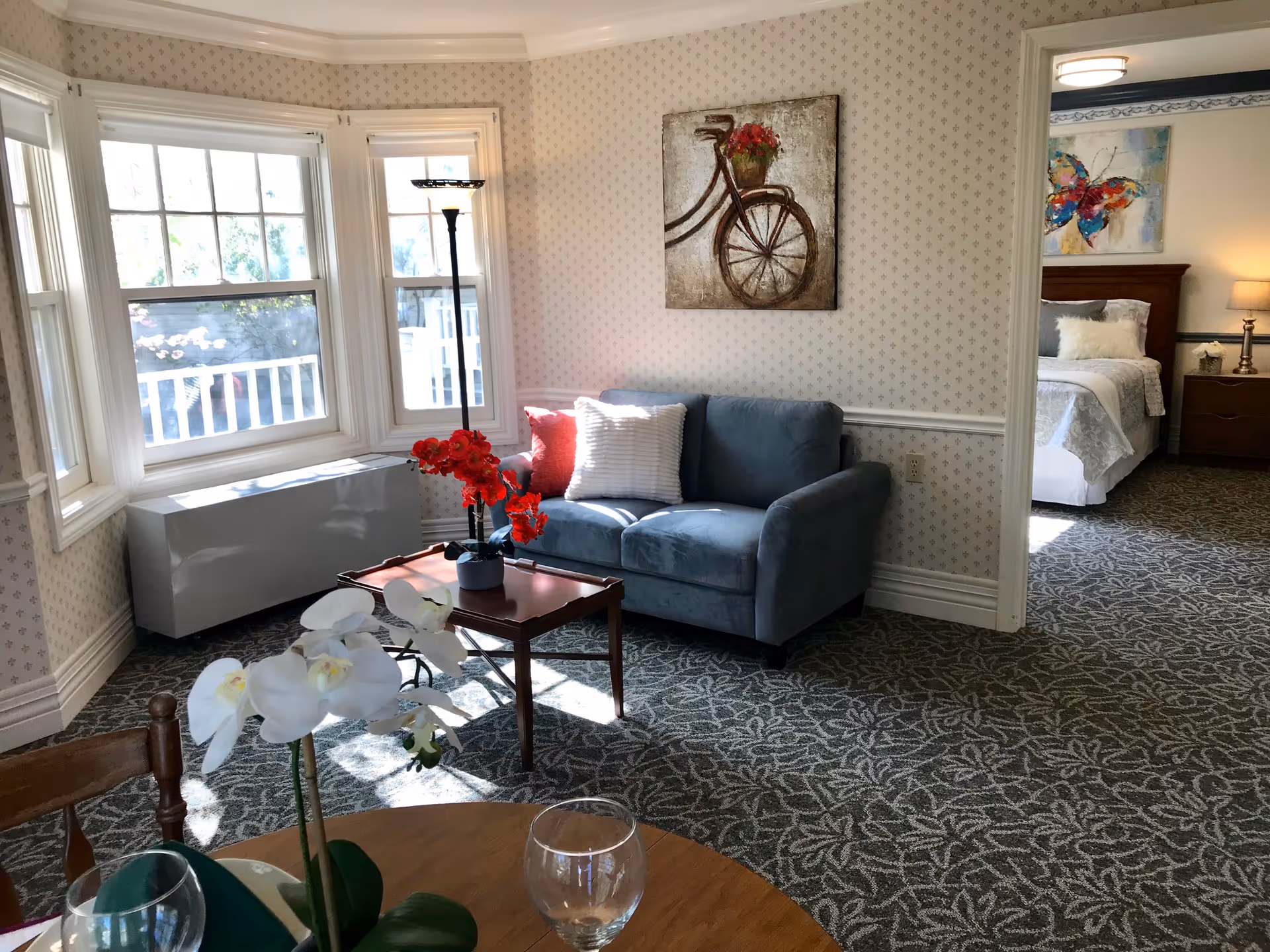 Sunlit living room with a blue loveseat, small wooden coffee table holding red flowers, bay windows, and a view into an adjacent bedroom.