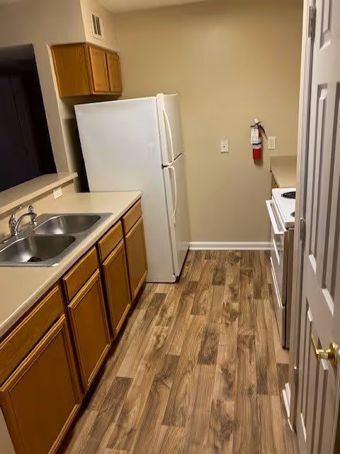 A small kitchen with wooden cabinets, a double sink, a white refrigerator, a white stove, and wood-patterned flooring. A fire extinguisher is mounted on the wall near the stove.