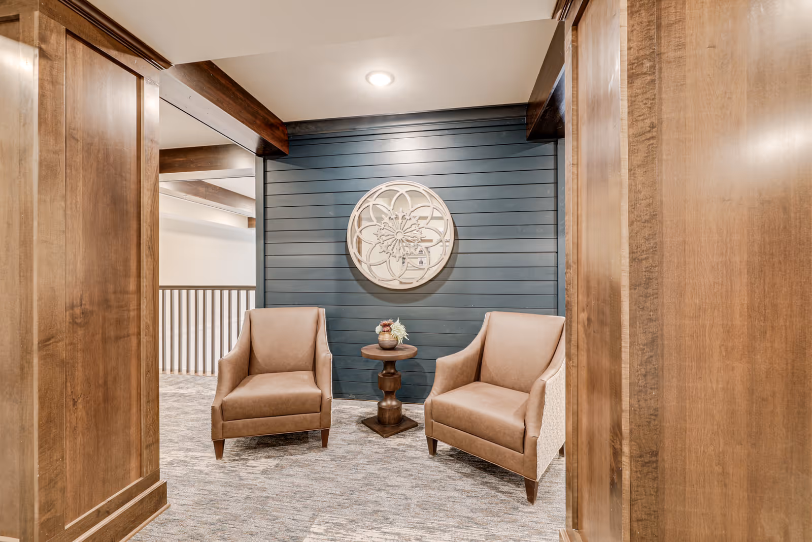 A cozy seating area with two brown armchairs facing each other and a small round wooden table with a decorative vase in between. The background features a dark blue shiplap wall with a large circular decorative wall art. The space is framed by wooden beams and panels, with carpeted flooring and a ceiling light above.