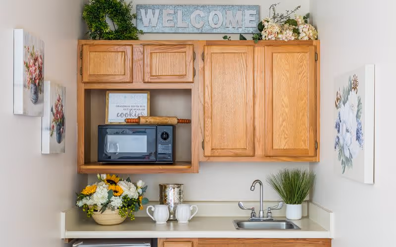 A small kitchen area with wooden cabinets, a microwave, a rolling pin, and decorative flowers. There is a sign that says 'WELCOME' on top of the cabinets and a framed quote about cookies inside the cabinet. The countertop has two white mugs, a silver container, and a small potted plant next to a sink with a faucet. Floral paintings are hung on the walls on either side.