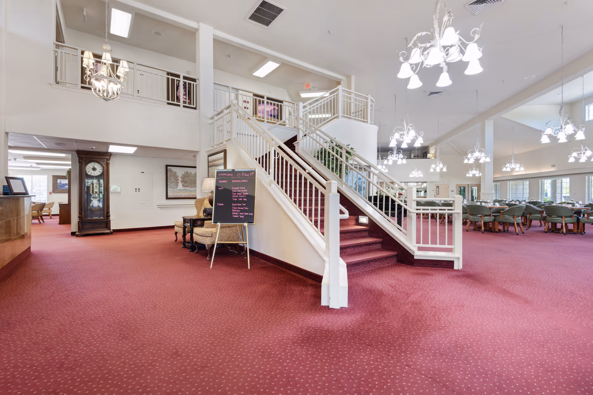 Interior view of a senior living facility featuring a central staircase with white railings and red carpet. The space has multiple chandeliers hanging from the ceiling, a large clock against the wall, seating areas with chairs and tables, and a menu board near the staircase. The area is spacious with red carpeting and large windows allowing natural light.