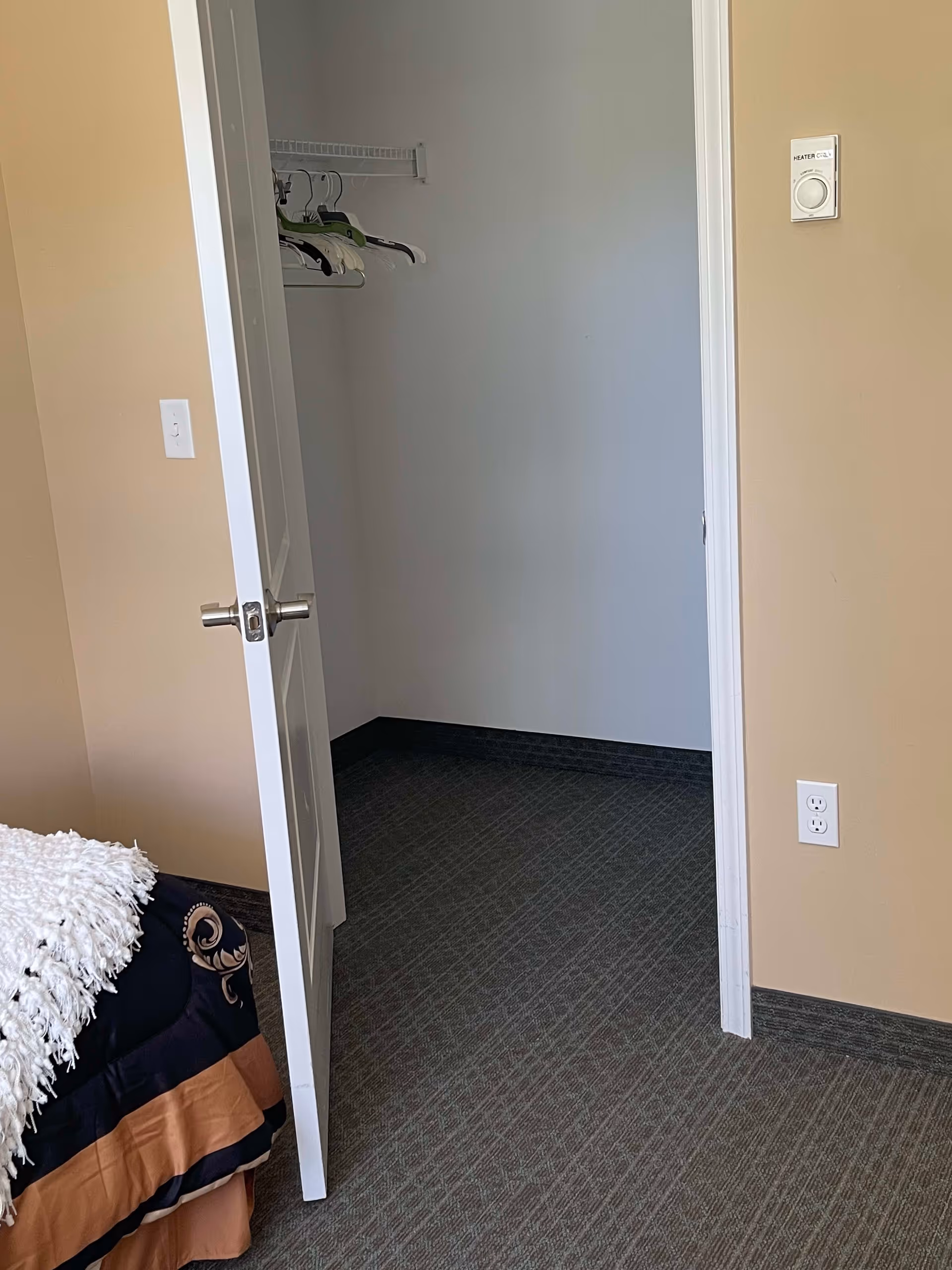 View of a partially open white door leading to an empty closet with a few hangers on a wire shelf. The room has beige walls, a bed with a black and brown bedspread and a white textured throw blanket, and a carpeted floor.