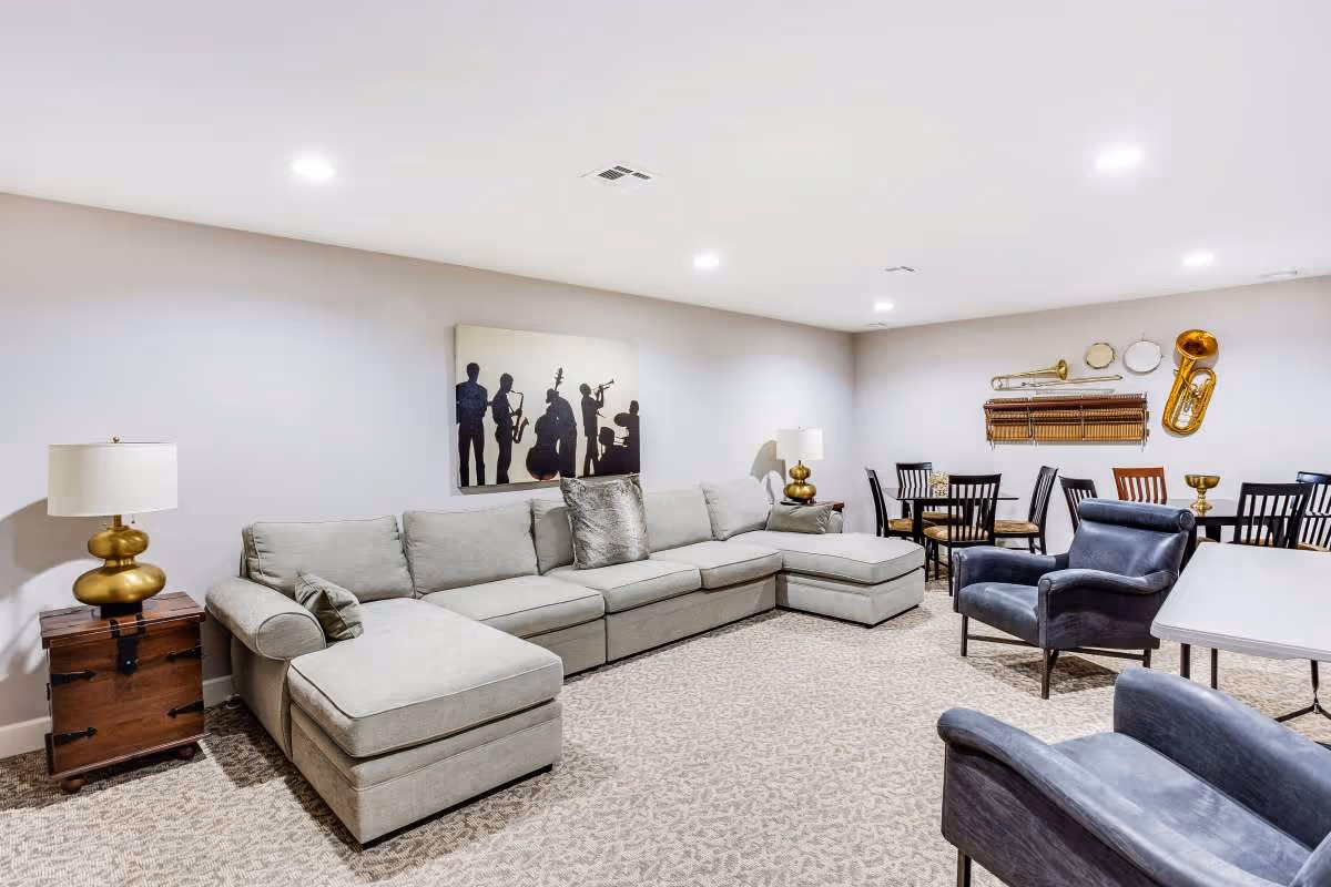 A spacious living room with a large light gray sectional sofa, two blue armchairs, and a wooden side table with a gold lamp. The walls are decorated with a silhouette painting of musicians and various brass musical instruments mounted on the wall. There are two dining tables with black chairs in the background, and the room is well-lit with recessed ceiling lights.
