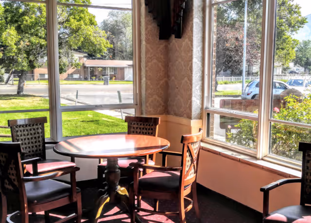 A sunlit corner of a room with large windows showing a view of trees, grass, and parked cars outside. Inside, there is a round wooden table surrounded by four wooden chairs with cushioned seats and patterned backs. The walls have a light-colored wallpaper with a subtle pattern, and the floor is carpeted in a dark red color.