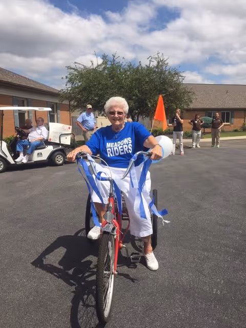 An elderly woman wearing a blue Meadows Riders t-shirt and white pants is riding a red tricycle decorated with blue and white streamers on the handlebars. She is smiling and outdoors on a paved area with a building and several people in the background, including a person sitting in a golf cart.
