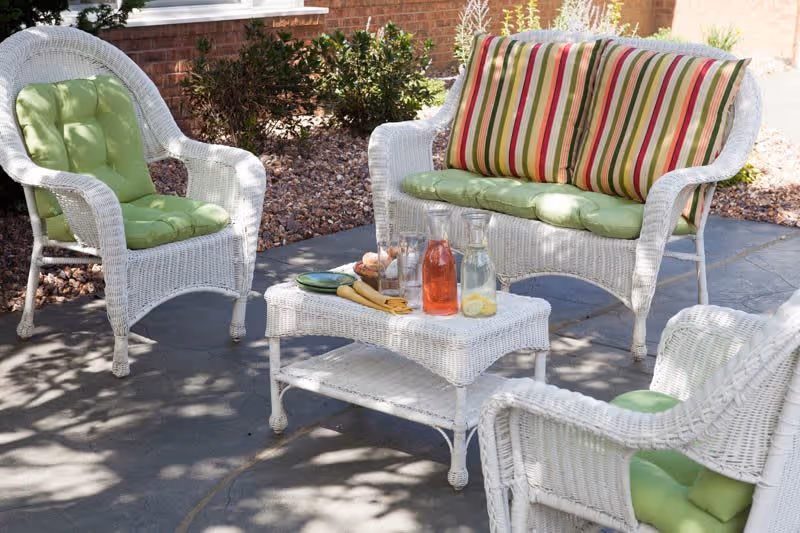 Outdoor patio area with white wicker furniture including two armchairs and a loveseat with green and striped cushions. A matching wicker coffee table holds two glass pitchers with beverages and some plates. The patio is surrounded by landscaping with rocks and plants.