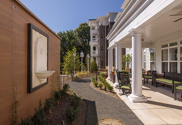 Landscaped outdoor walkway and patio with white columns, benches, greenery, and a wall-mounted fountain beside a senior living building.