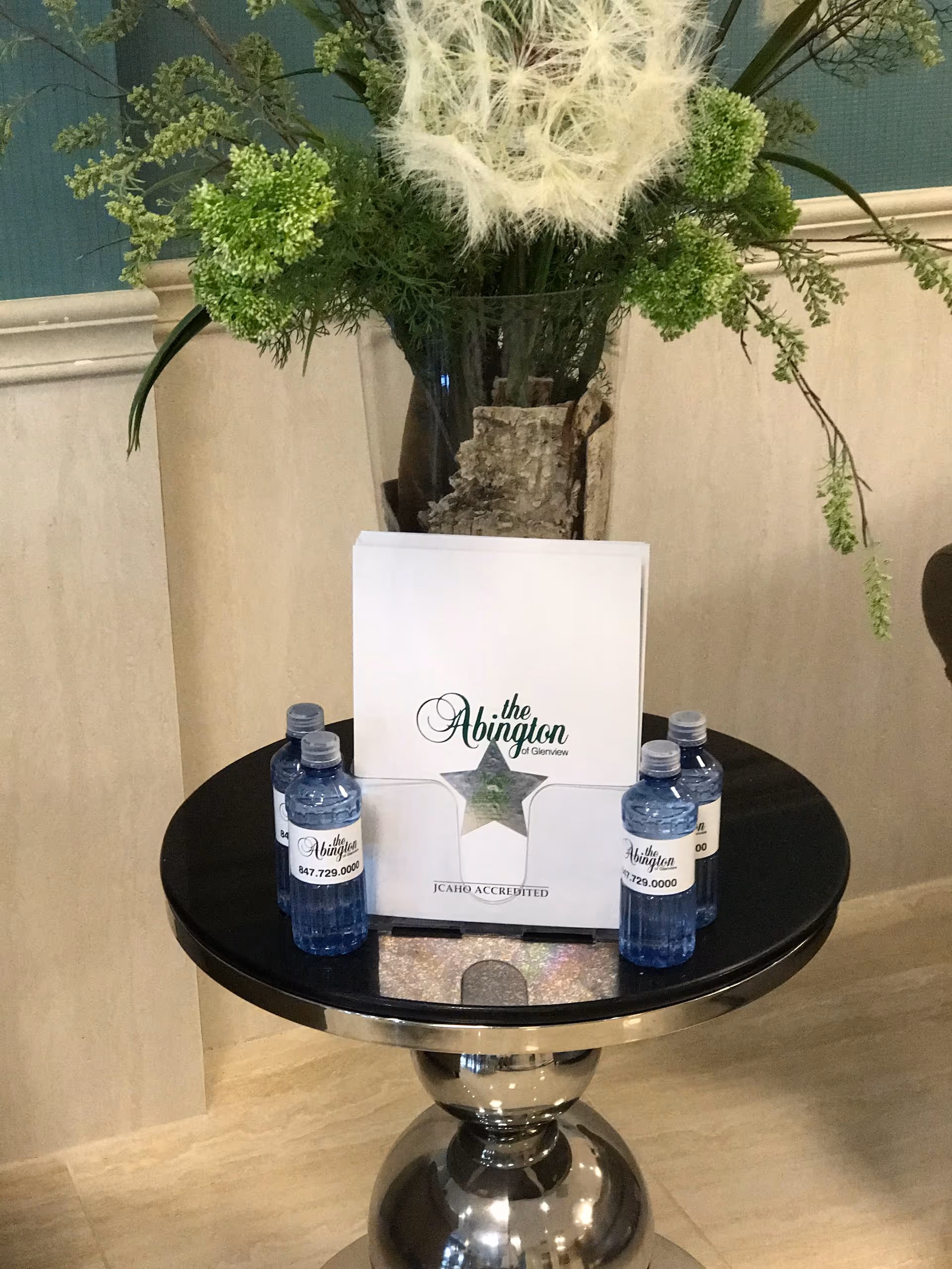 A round pedestal table in a lobby with a vase of flowers, a brochure for "the Abington", and four bottled water bottles.