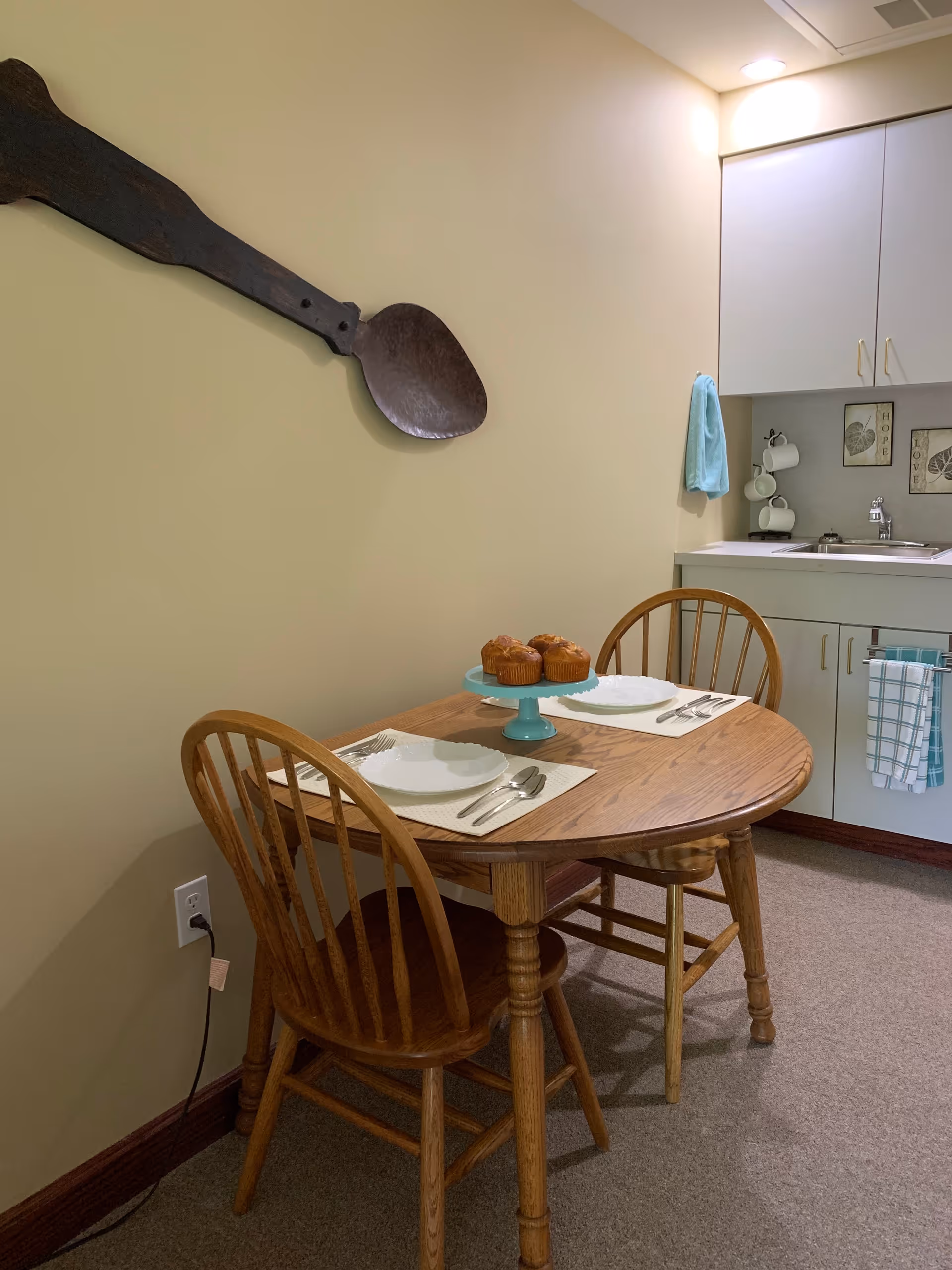 A small dining area with a wooden table set for two with white plates, silverware, and placemats. On the table is a blue cake stand holding three muffins. Two wooden chairs are placed at the table. In the background, there is a kitchenette with white cabinets, a sink, a towel hanging on the cabinet handle, and a blue hand towel hanging on the wall. A large decorative wooden spoon is mounted on the beige wall above the table.