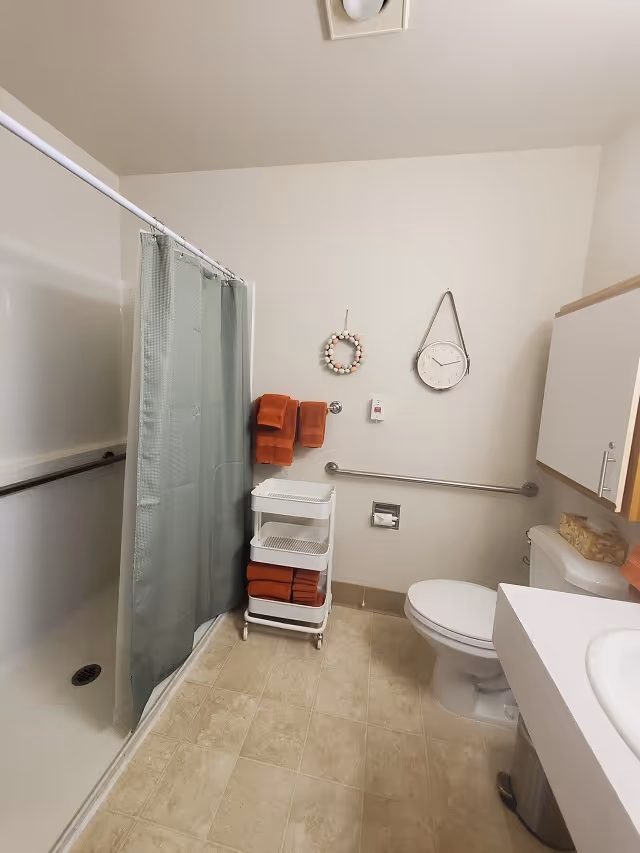 Accessible bathroom with a shower and curtain, toilet, grab bars, orange towels, wall clock, and a rolling storage cart.