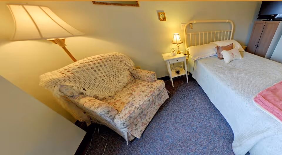 A cozy bedroom featuring a bed with white bedding and decorative pillows, a floral upholstered armchair with a crocheted throw draped over it, a small white nightstand with a lamp and clock, and a wooden cabinet with a TV on top.