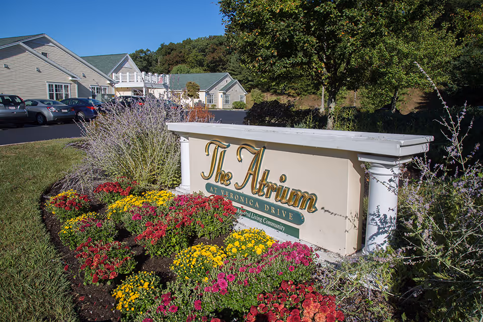 Outdoor view of the entrance sign for The Atrium at Veronica Drive, surrounded by colorful flowers and greenery, with the facility buildings and parked cars visible in the background under a clear blue sky.
